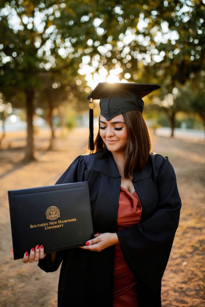A graduate in a cap and gown holds a diploma from Southern New Hampshire University outdoors at sunset.