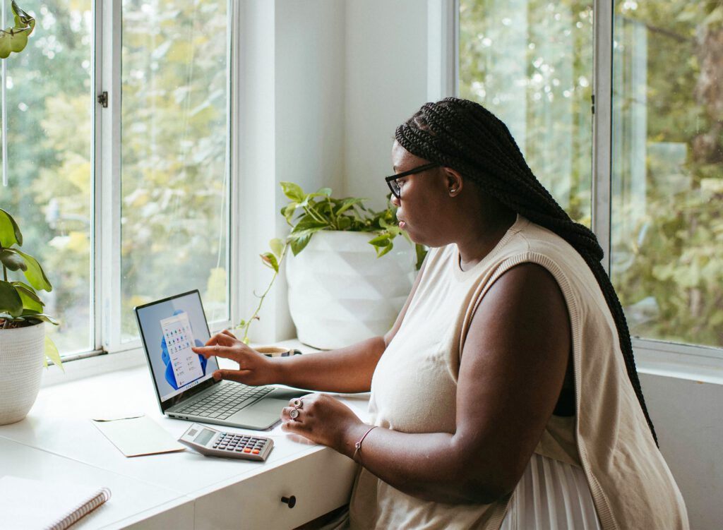 A woman with braided hair and glasses sits at a desk by a window, using a laptop with a calculator and notebook beside her.